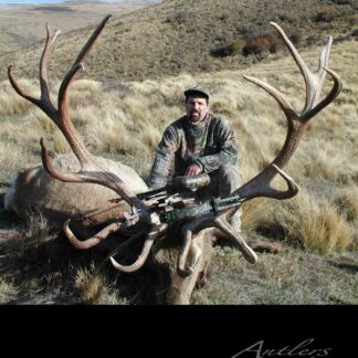 A man in camouflage kneels behind a Rudzinis Elk with impressive antlers in a grassy, hilly landscape. A crossbow rests on the elk, and there's an Antlers by Klaus watermark on the image.