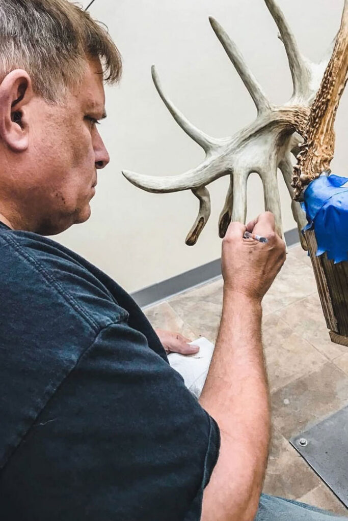 A man in a dark shirt carefully paints or restores a large antler, which is partially covered with blue tape, while seated indoors on a tiled floor.